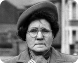Portrait of an elderly woman wearing a dark beret and glasses, looking at the camera outdoors.